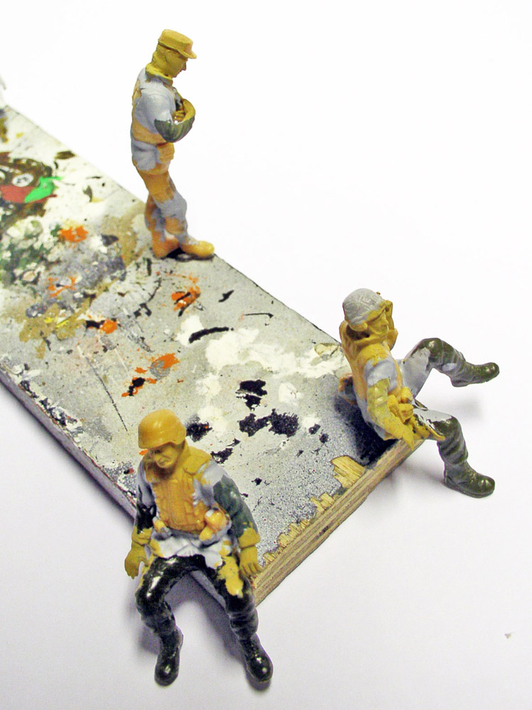 Trois figurines de soldats miniatures en cours de peinture posées sur une planche tachée