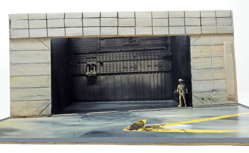 Diorama de hangar en pierre avec grande porte métallique sombre et figurine de pilote