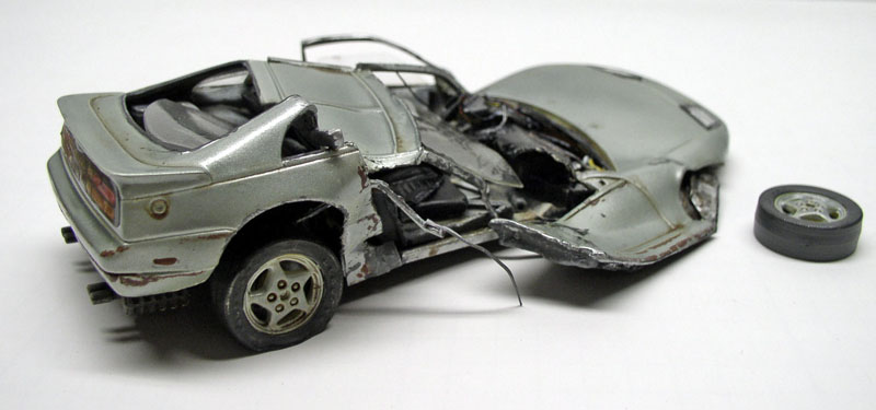 Maquette de voiture de sport grise accidentée, carrosserie déchirée et roue détachée posée à côté