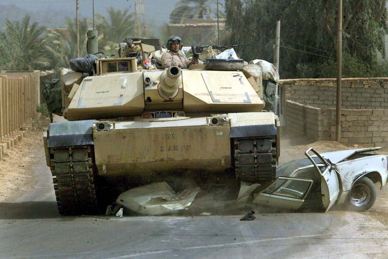 Char Abrams M1 A1 sable écrasant une voiture blanche sur une route poussiéreuse