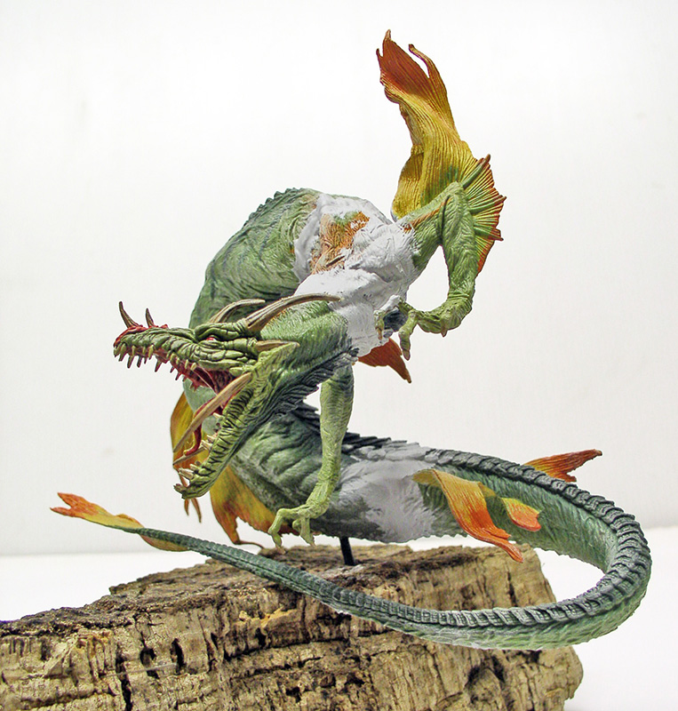 Maquette de créature aquatique verte aux nageoires orangées et longue queue, posée sur écorce