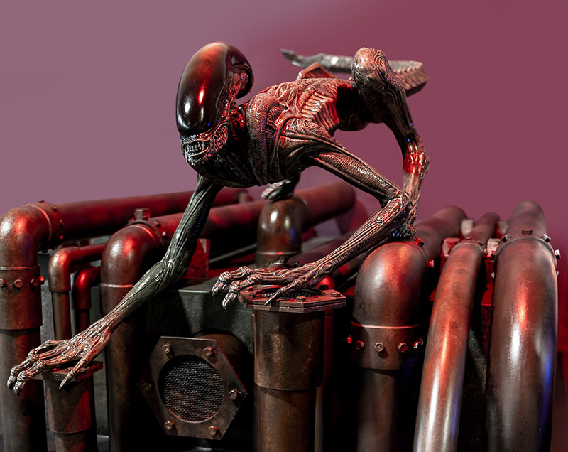 Maquette du xénomorphe d'Alien 3 accroupi sur des tuyaux métalliques rouillés devant un fond violet