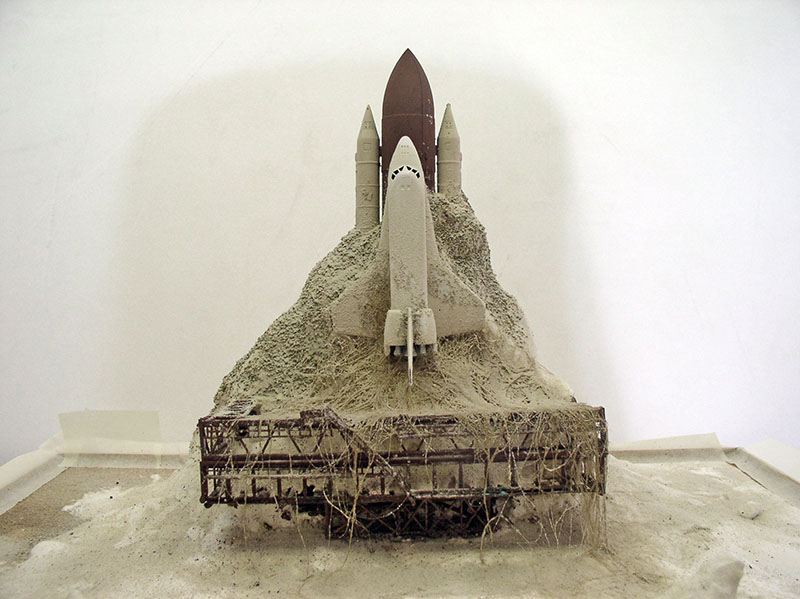 Maquette de navette spatiale avec boosters émergeant d'un relief sablonneux sur socle métallique rouillé