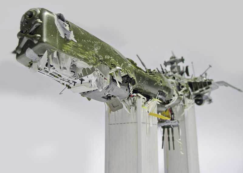 Maquette de l'Arcadia d'Albator représentée écrasée et disloquée, coque verte déchirée posée sur supports blancs
