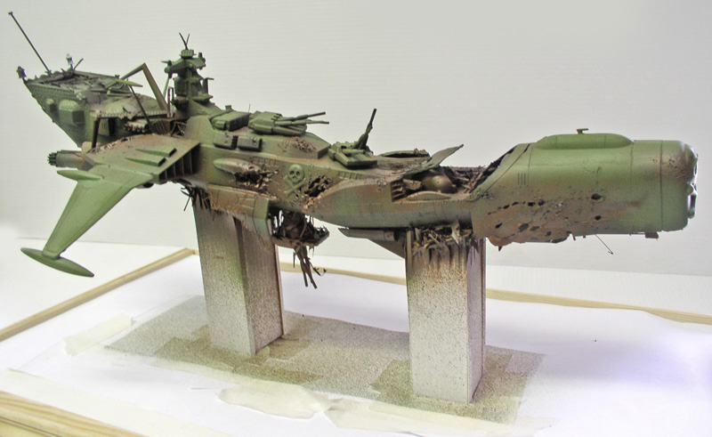 Maquette du cuirassé Arcadia vert vieilli posée sur deux piliers, montrant les dégâts du crash