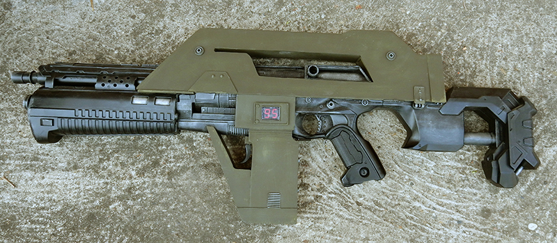 Réplique de fusil futuriste kaki pour cosplay avec compteur numérique rouge posée au sol