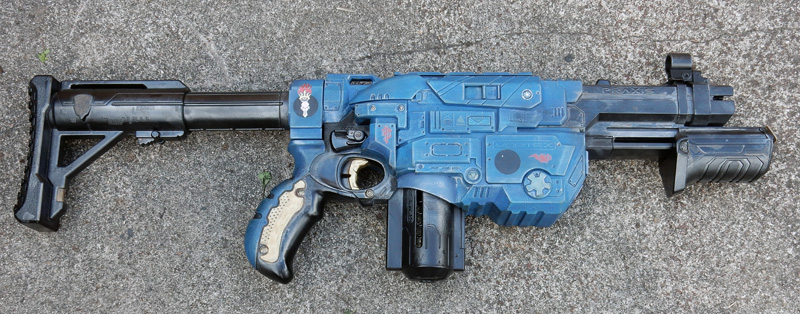 Réplique d'arme futuriste de cosplay peinte en bleu, posée sur béton gris