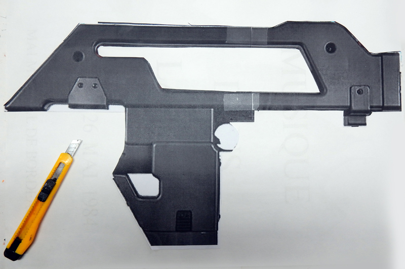 Silhouette de pistolet futuriste découpée dans du carton gris à côté d'un cutter jaune