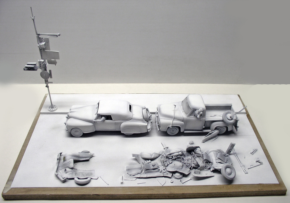Maquette grise en cours de montage avec deux voitures et pièces détachées éparpillées sur un socle