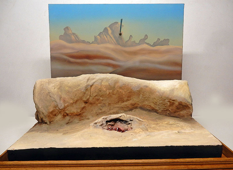 Diorama de maquette désertique avec rocher creusé et décor de fond peint évoquant un paysage aride