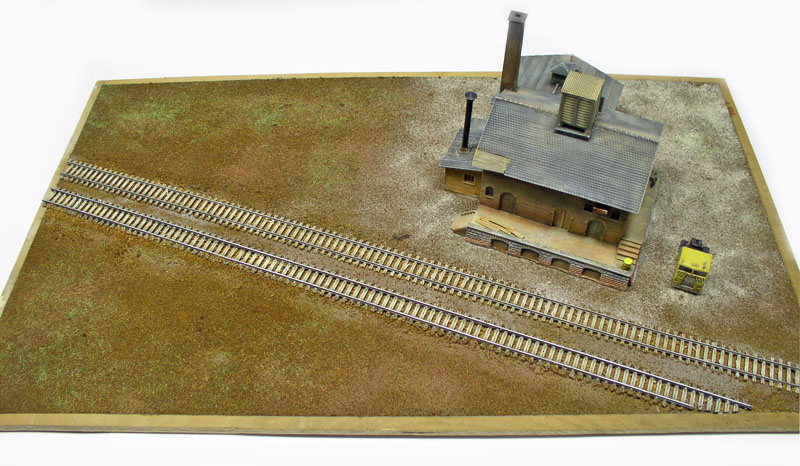 Maquette ferroviaire avec voies parallèles et bâtiment industriel en pierre sur décor de terrain
