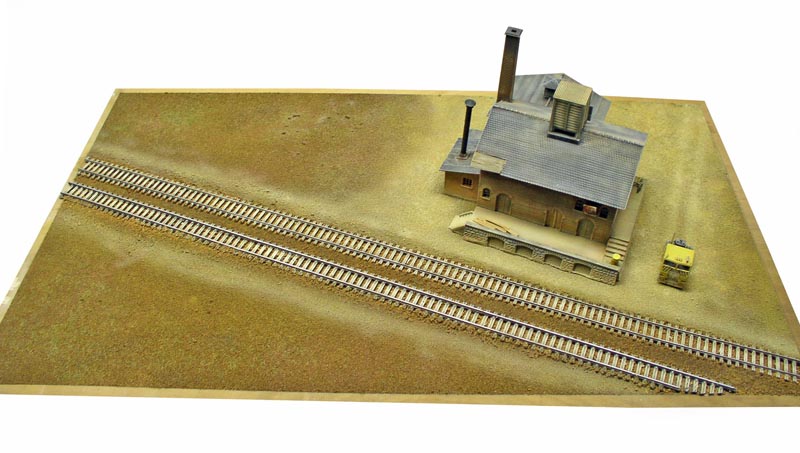 Maquette ferroviaire avec voies parallèles et bâtiment industriel à cheminée sur décor désertique