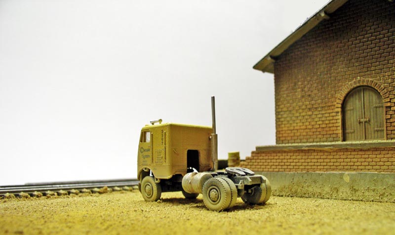 Maquette de tracteur routier jaune près d'un bâtiment en briques et voie ferrée