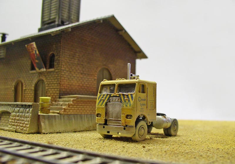 Tracteur routier miniature jaune patiné devant un bâtiment en briques près d'une voie ferrée