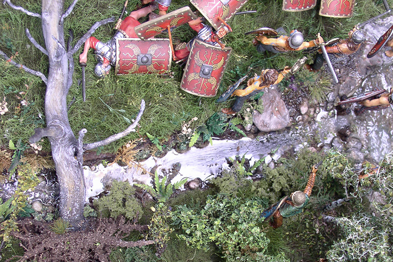 Légionnaires romains miniatures avec boucliers rouges progressant près d'un arbre dans un diorama végétalisé