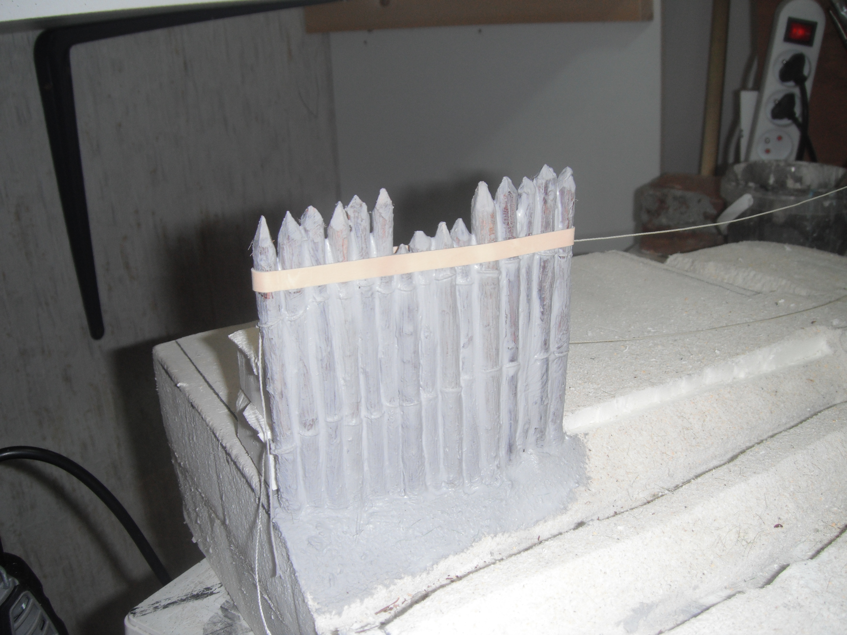 Palissade de pieux pointus en cours de fabrication maintenue par un élastique sur l'établi