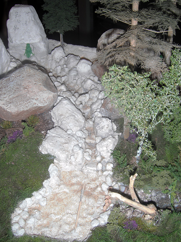 Maquette d'un sentier rocheux blanc serpentant entre arbres miniatures et végétation, décor du camp romain