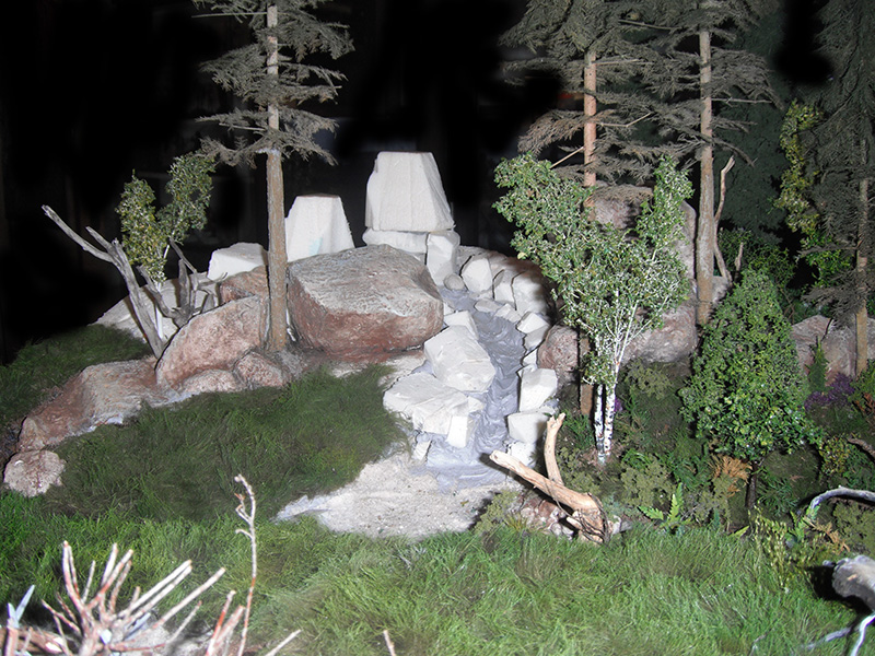 Maquette de diorama forestier avec rochers, arbres miniatures et ruisseau en construction pour le Camp Romain