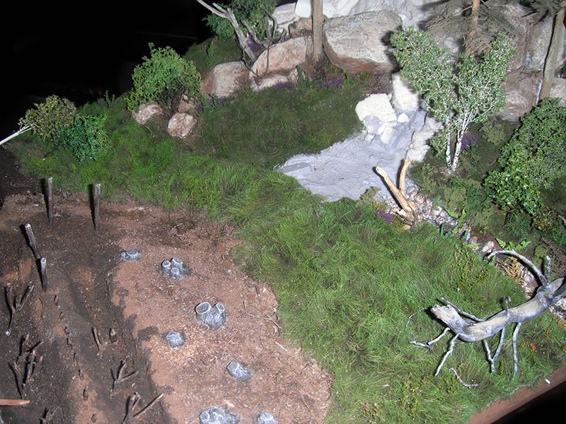Diorama de camp romain avec talus de terre, pieux défensifs, herbe, rochers et arbres miniatures