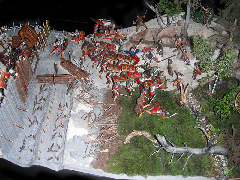 Diorama de bataille opposant légionnaires romains aux boucliers rouges et guerriers barbares devant un rempart fortifié