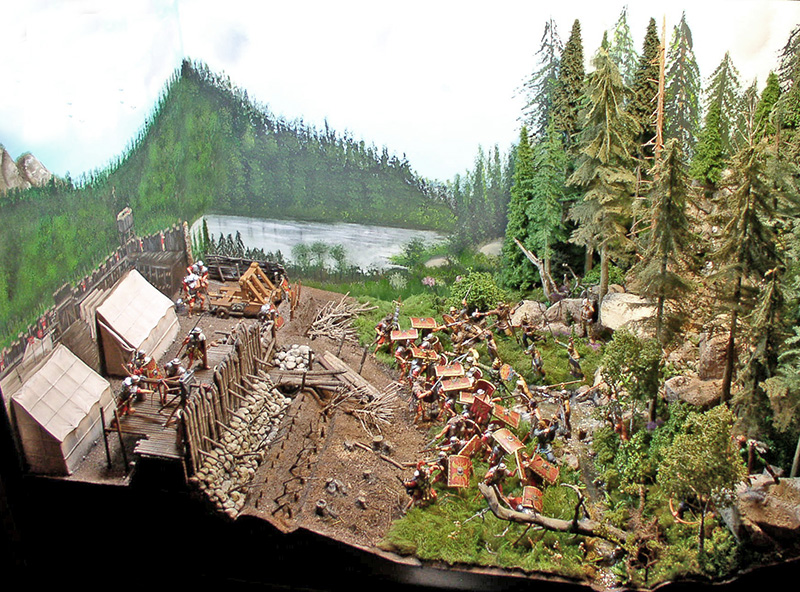 Diorama d'un camp romain fortifié assiégé par des soldats près d'un lac forestier