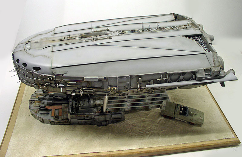 Maquette de cargo spatial gris détaillé posé sur un diorama sablonneux avec petit véhicule terrestre