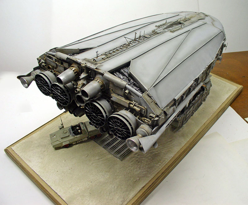 Maquette de cargo spatial gris avec moteurs arrière et petit véhicule sur socle