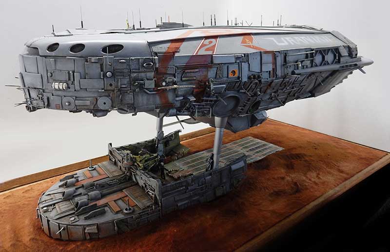 Maquette de cargo spatial gris sur socle, surplombant une plateforme d'atterrissage détaillée