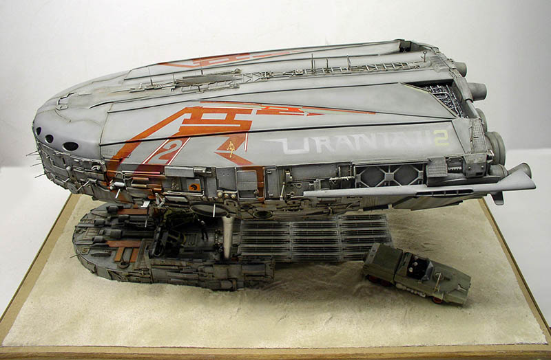 Maquette de cargo spatial gris vieilli posé sur diorama sablonneux avec petit véhicule terrestre