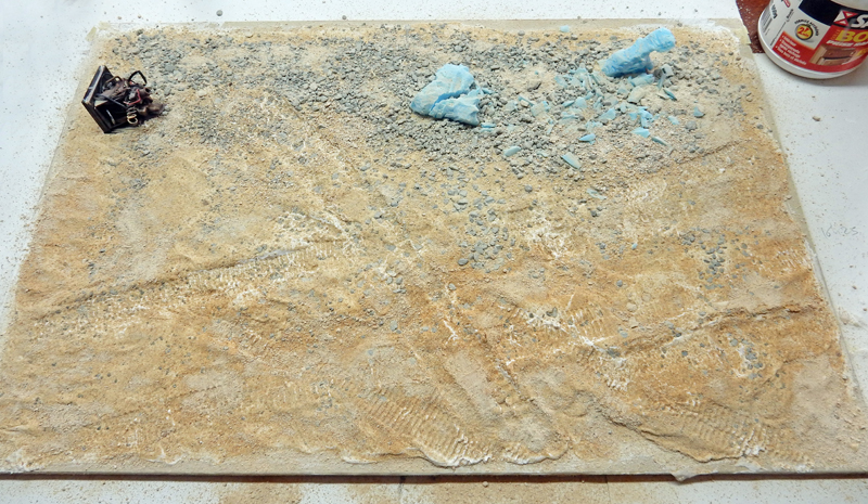 Socle de diorama en cours avec sol sablonneux texturé, débris bleus et petite figurine