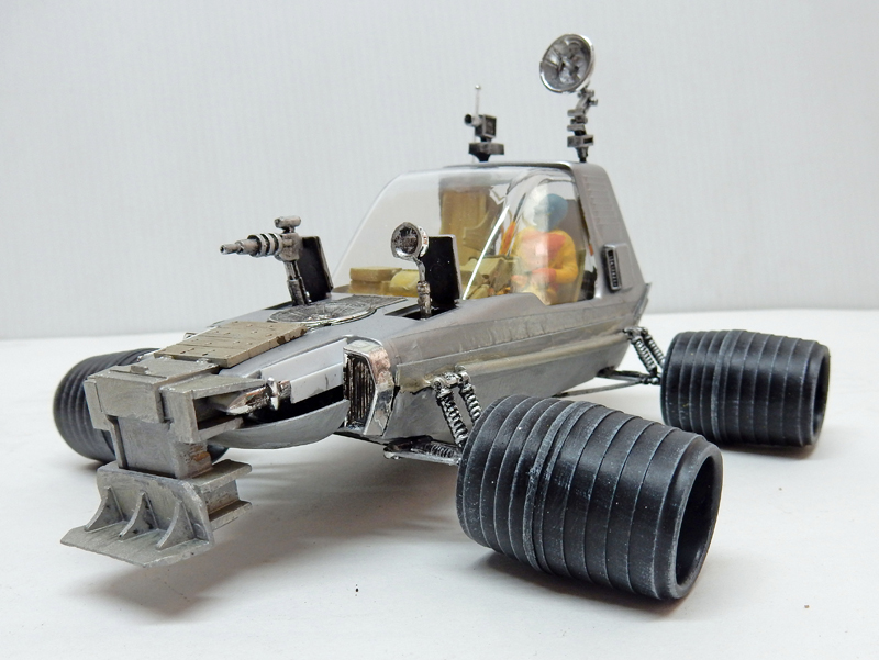 Maquette de véhicule lunaire argenté à cockpit transparent et roues cylindriques, sur fond clair