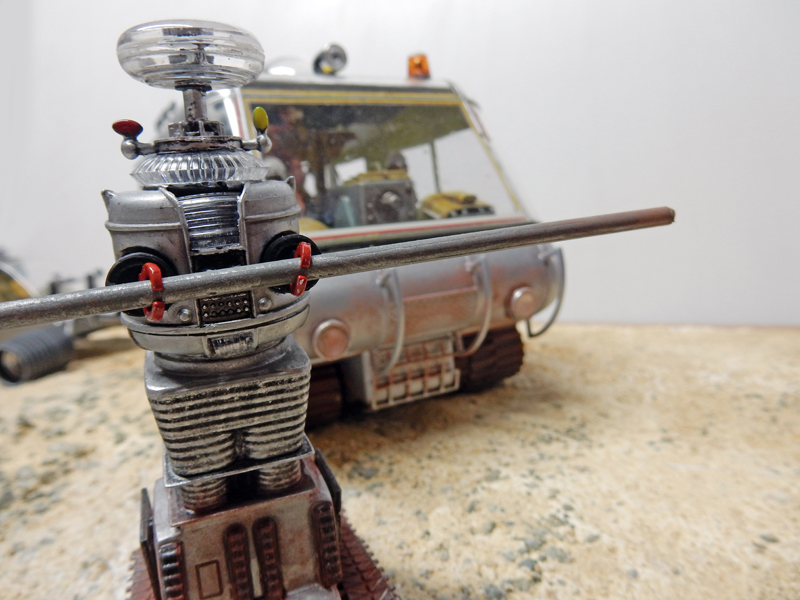 Maquette de robot rétro patiné devant un véhicule chenillé argenté dans un diorama