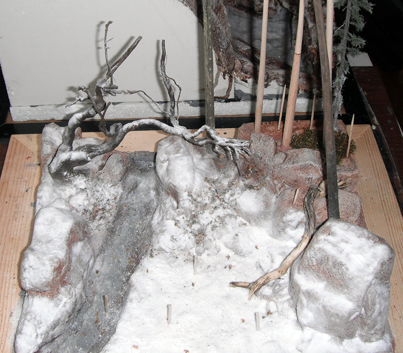 Diorama enneigé en construction avec rochers, arbres morts et piquets sur socle bois