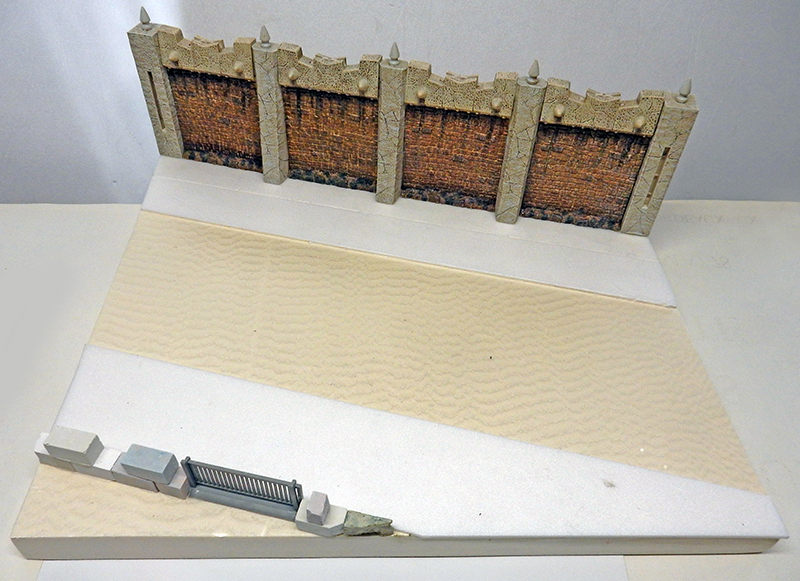 Maquette de mur en briques crénelé sur socle de polystyrène avec terrain sablonneux et grille en construction