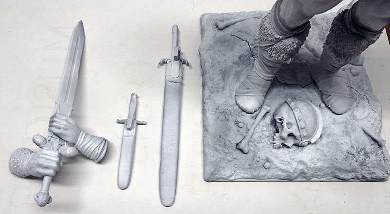 Pièces de maquette non peintes : épées, main et socle rocheux orné d'un crâne casqué