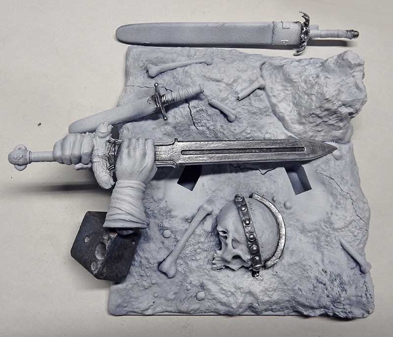 Pièces de maquette non peintes : épées, mains, crâne et ossements sur socle rocheux