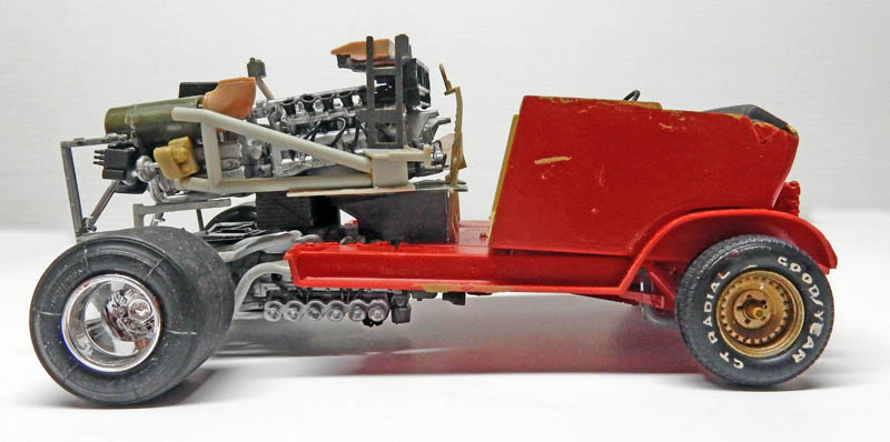 Maquette de hot rod rouge en construction, profil montrant châssis, gros moteur et pneus larges
