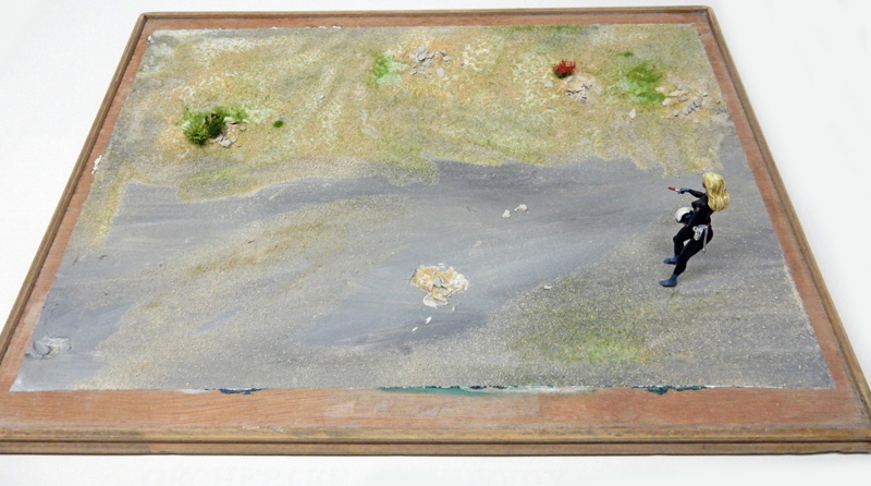 Diorama de maquettisme avec sol désertique, touffes de végétation et figurine féminine armée en noir