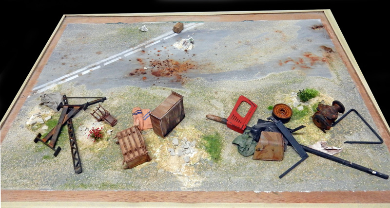 Diorama de maquette montrant un terrain désolé avec route, caisses, débris rouillés et objets abandonnés