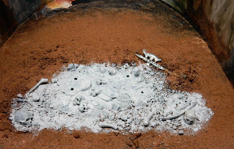 Crânes et ossements gris sur sol de terre ocre avec hache dans un diorama