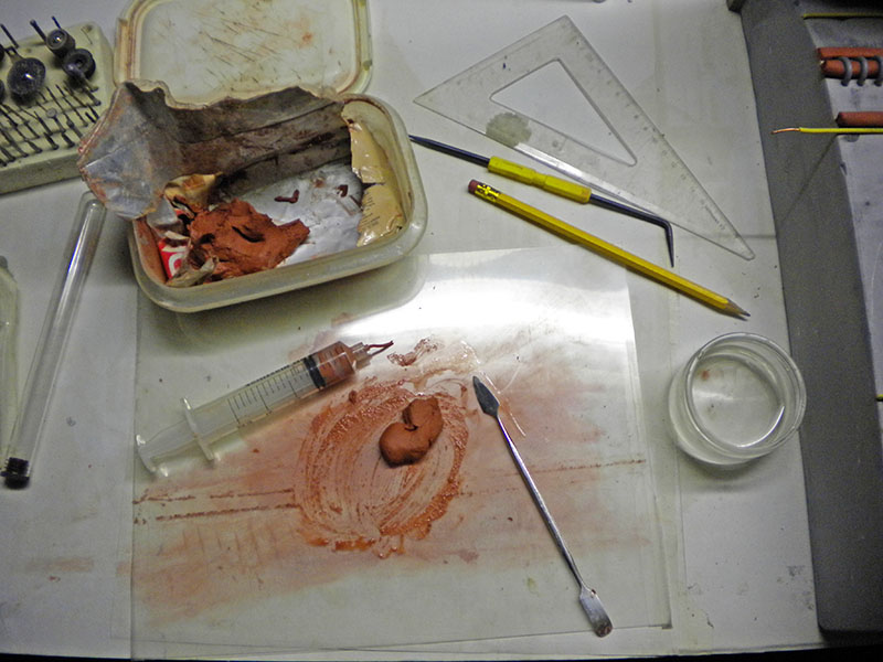 Plan de travail avec pâte à modeler ocre, seringue, spatule, crayon et équerre pour décor miniature