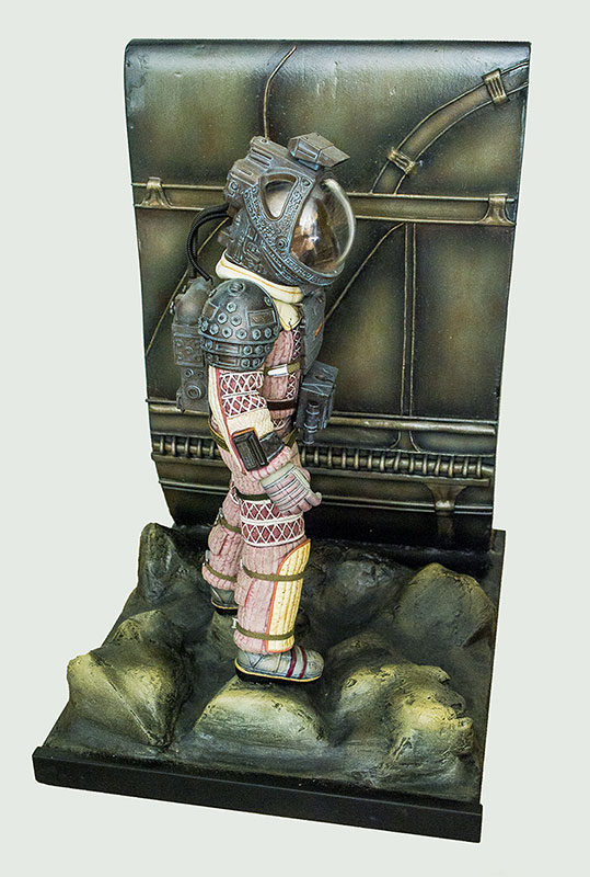 Figurine d'astronaute en scaphandre devant un décor mural mécanique sur sol rocheux