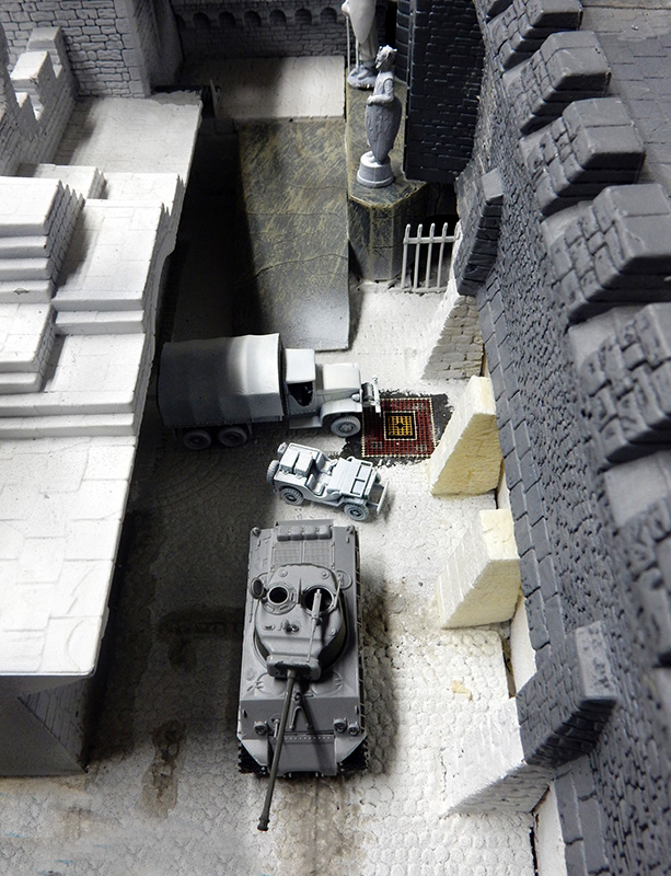 Maquette grise en cours : char, jeep et camion dans une ruelle fortifiée