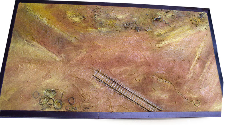 Maquette de diorama avec sol désertique ocre, rails miniatures et petits éléments métalliques posés dessus