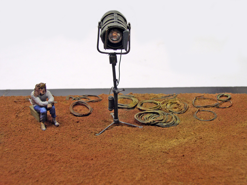 Maquette de scène de tournage avec projecteur sur trépied, figurine assise et câbles enroulés au sol