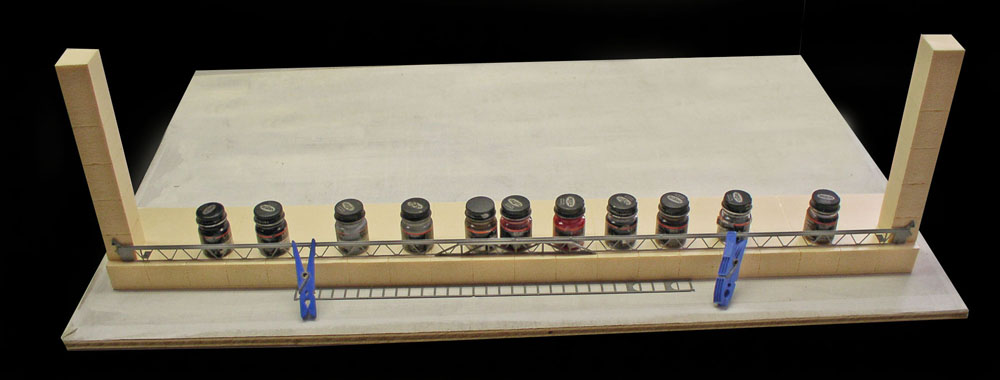 Maquette de pont métallique en cours de collage, lesté par des pots de peinture et pinces à linge