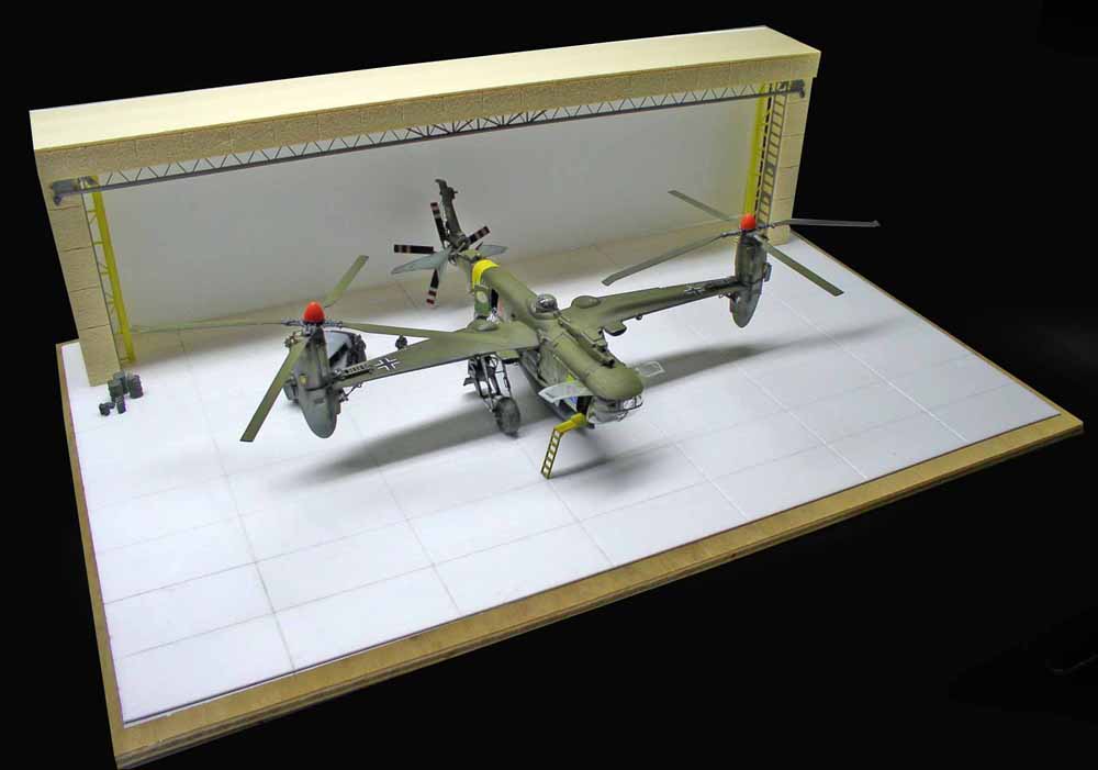 Maquette d'hélicoptère vert militaire à deux rotors devant un hangar sur diorama