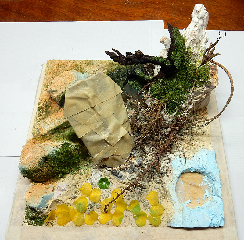 Maquette de diorama en cours avec rochers, mousse, racines et fleurs jaunes pour décor de dragon