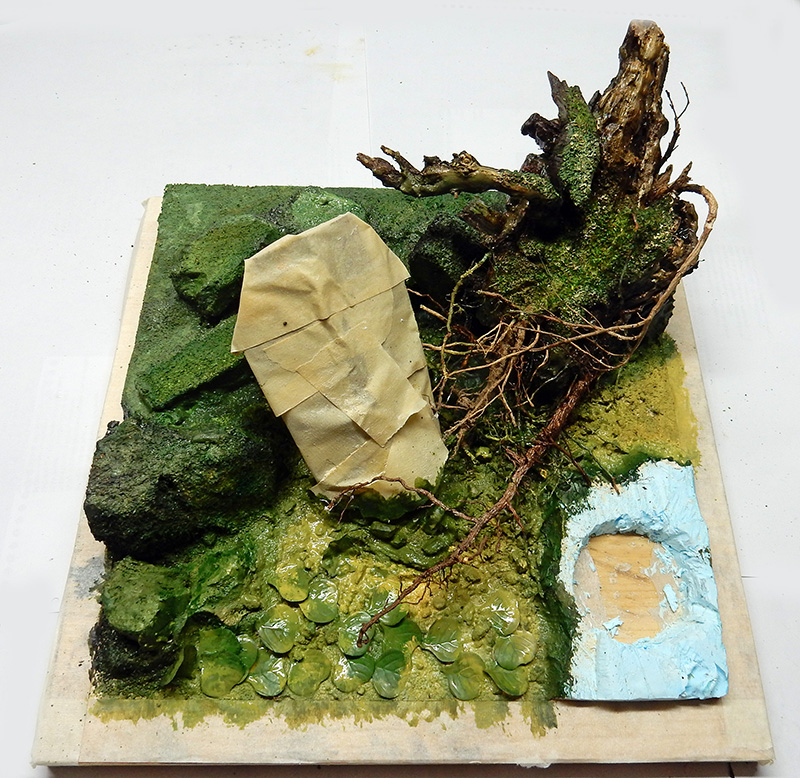 Diorama de maquette en cours avec mousse végétale, racines, rocher et plan d'eau bleu