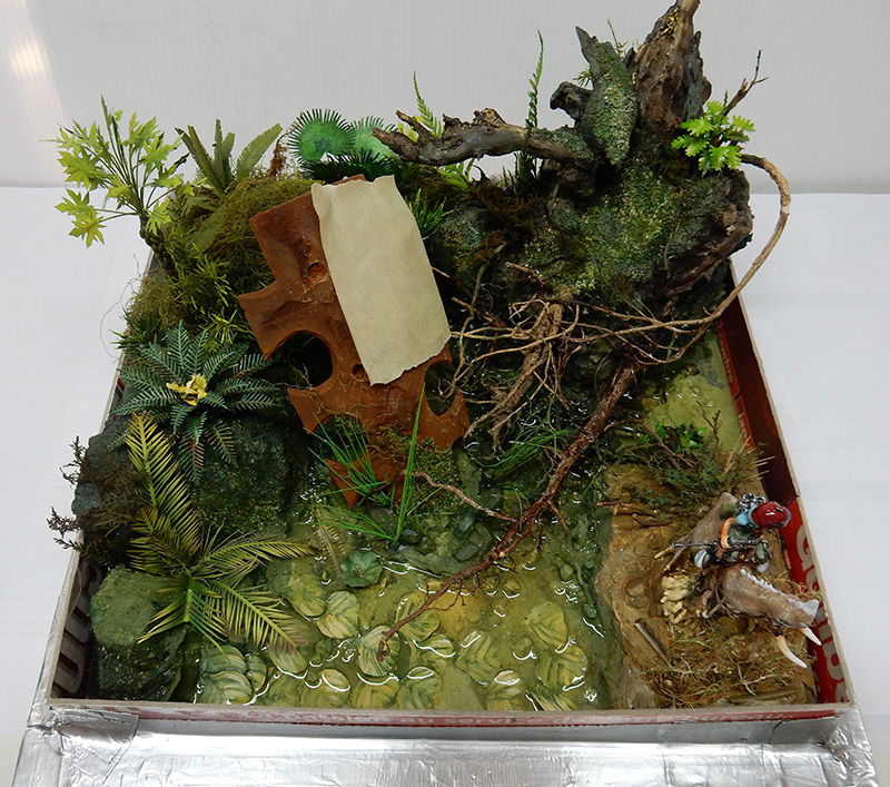 Diorama de maquettisme représentant un marécage végétalisé avec dragon vert perché et figurine